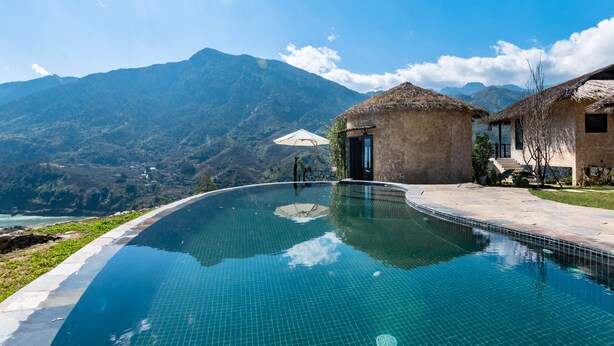 Una piscina con vista a la montaña, un edificio con techo de paja y un cielo despejado.