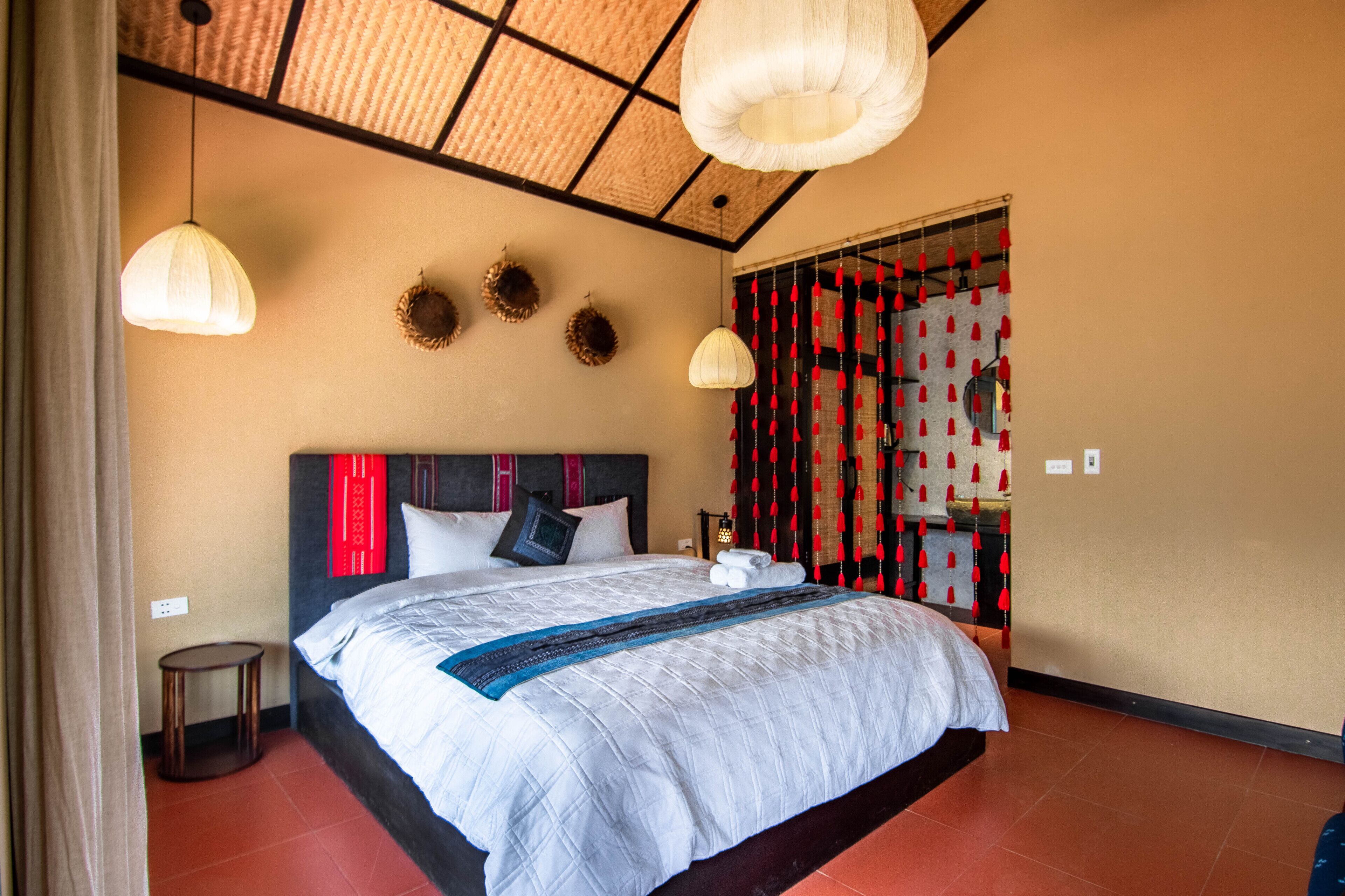 A neatly made bed with a white comforter and a blue accent, a round side table, a woven ceiling, and a red and black curtain with tassels.