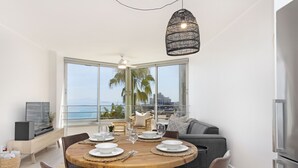 A modern dining area with a round wooden table set for four, a large window with a view of palm trees and the ocean, and a ceiling fan. - Welgelegen 608 (Cape Town)