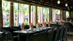 An outdoor dining area with wicker tables and chairs, large windows with a view of palm trees, and a covered roof structure.