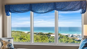 A bedroom with a large window overlooking a coastal view, a bed with a patterned pillow, and a blue curtain. - Summit at Tops`l 810 - Beautiful Gulf Views! (Miramar Beach)
