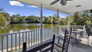 Um deck coberto com mesa e cadeiras, com vista para um lago tranquilo e cercado por árvores.