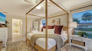 A four-poster canopy bed with a woven throw blanket, a bedside table with a lamp, and a large window with a view of palm trees and a building. - Open Labor Day Weekend |HTD Pool | 6 TVS | 2 King beds (Bradenton)