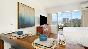 A hotel room with a large bed, a television, a dining area with a table, and a view of the city through large windows.
