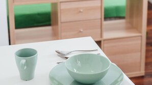 A room with a white table, a green mug, and a bowl on a matching plate. There is a wooden cabinet with multiple compartments and a green bed in the background.