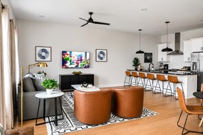 A modern living room with a sofa, coffee table, and ottomans, a dining area with bar stools, and a kitchen with a central island. - 2 Nashville Homes • Hot Tubs • Sleeps Up to 24 (Nashville)
