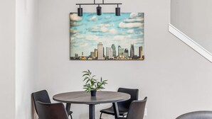 A dining area with a round table, four chairs, a potted plant, and a cityscape painting on the wall. - 2 Nashville Homes • Hot Tubs • Sleeps Up to 24 (Nashville)