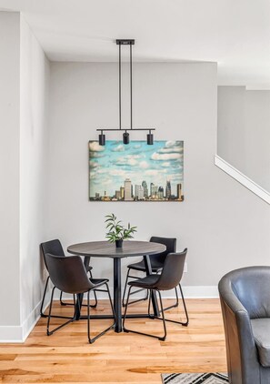 A dining area with a round table, four chairs, a potted plant, and a cityscape painting on the wall.