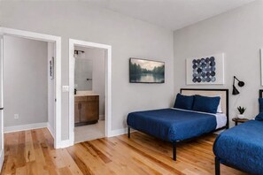 A bedroom with two beds, a wooden floor, a bathroom with a mirror, and a painting on the wall.