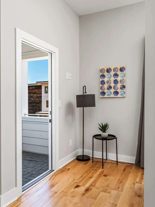 A hallway with a wooden floor, a door leading outside, a wall-mounted lamp, and a small table with a plant.