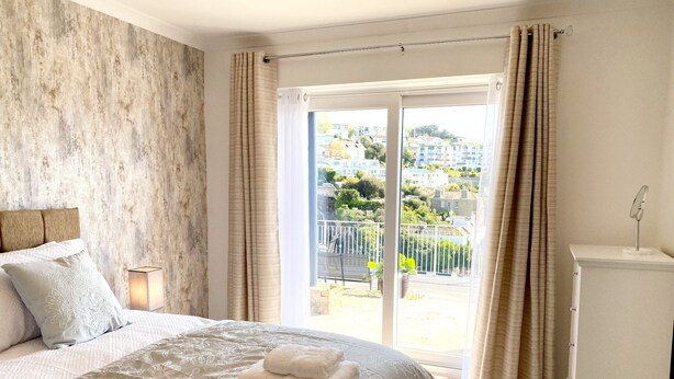 A bedroom with a large bed, a bedside table with a lamp, a window with curtains, and a view of a balcony with plants.