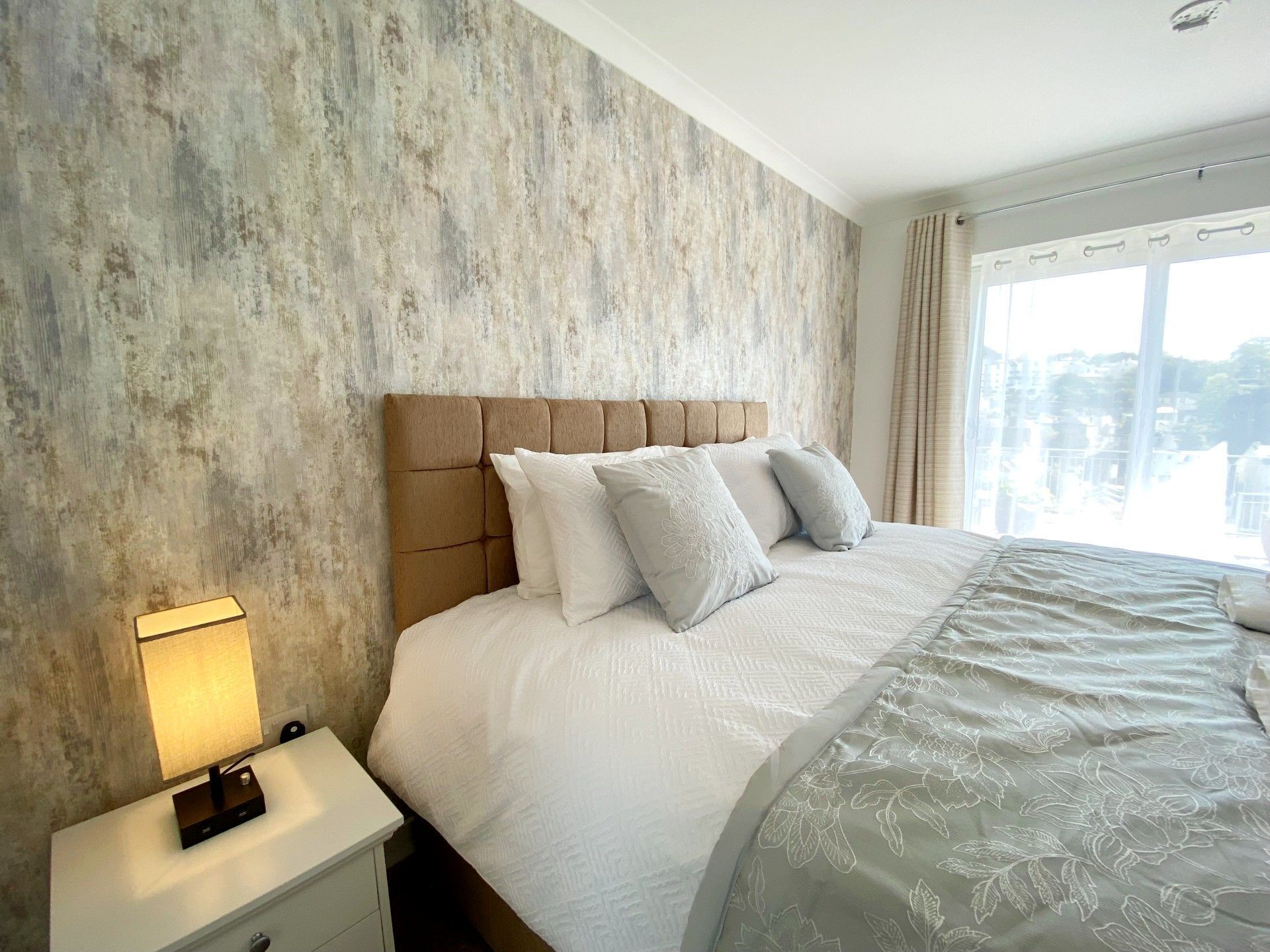 A hotel room with a large bed, a nightstand with a lamp, and a view of the city through the window.