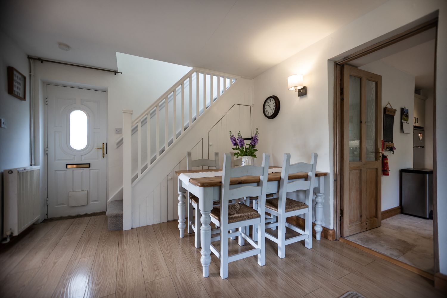 A dining area with a wooden table and white chairs, a staircase, and a clock on the wall.