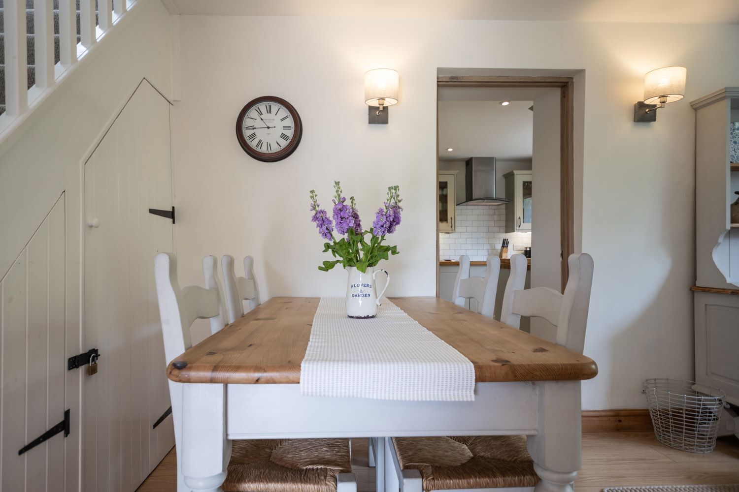 Une salle à manger avec une table en bois, des chaises blanches, un vase rempli de fleurs violettes et une horloge murale.