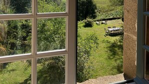 Vista a través de una ventana con cielo despejado y un paisaje cubierto de pasto, árboles, una mesa y sillas afuera.