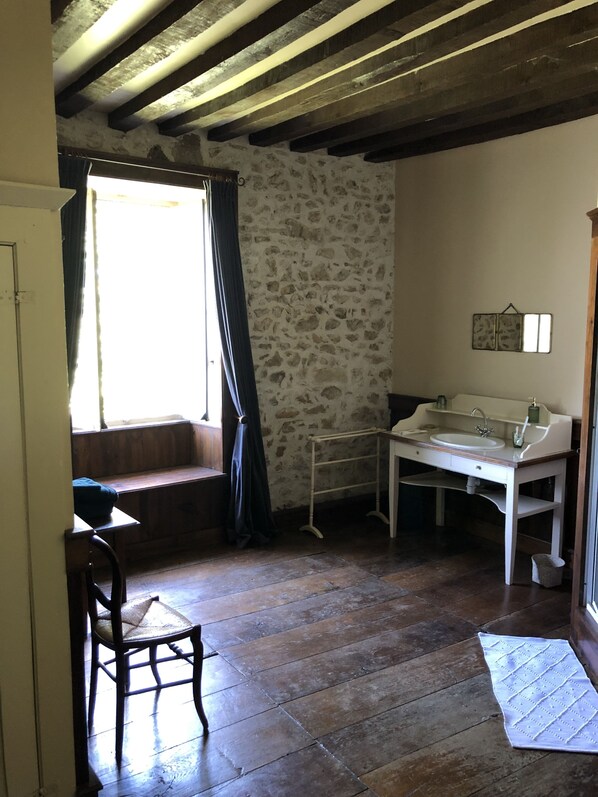 Une pièce rustique avec un mur en pierre, un plancher en bois, un banc, une chaise, une petite table avec un évier et une fenêtre avec des rideaux.