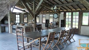 Un comedor rústico con una larga mesa de madera y sillas, una zona de cocina con estufa y lavaplatos, y ventanas amplias que ofrecen luz natural.