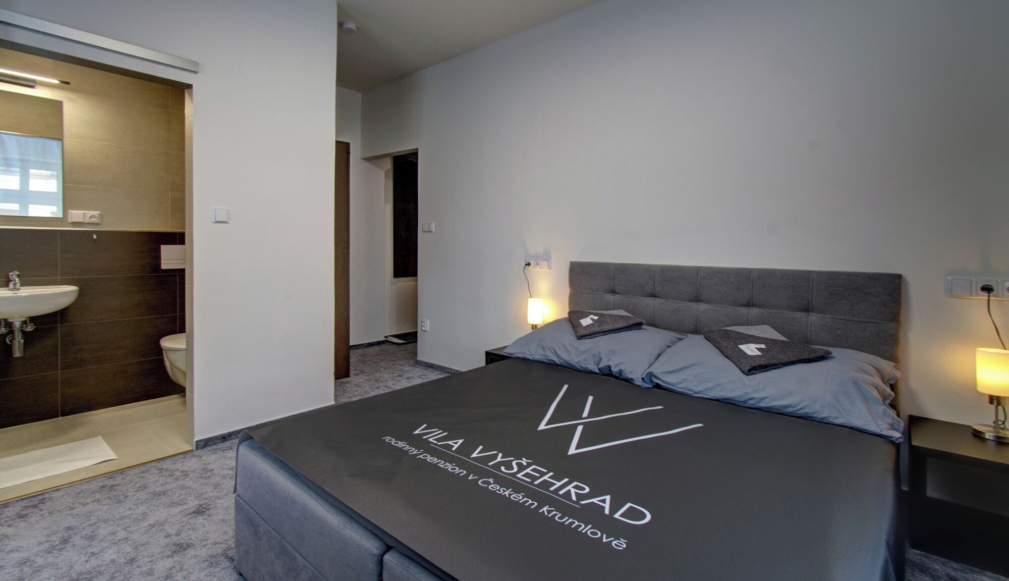 A modern hotel room with a large bed, two bedside lamps, a headboard, and a visible bathroom with a sink and toilet.