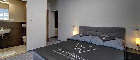 A modern hotel room with a large bed, two bedside lamps, a headboard, and a visible bathroom with a sink and toilet.