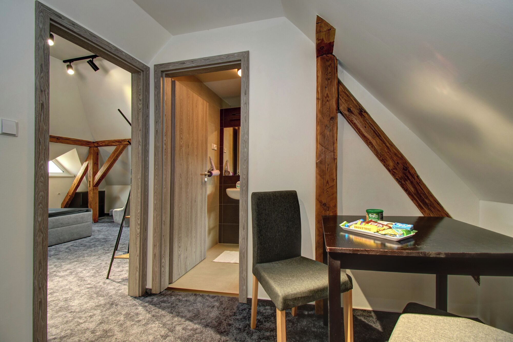 A loft-style room with exposed wooden beams, a gray chair, a small table with snacks, and a view into the bathroom.