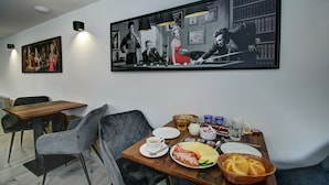 Un coin repas avec une table mise pour deux, où l’on aperçoit un repas composé de pain, de fromage et de jambon. Sur la table, on trouve deux chaises, une tasse à café et une théière.