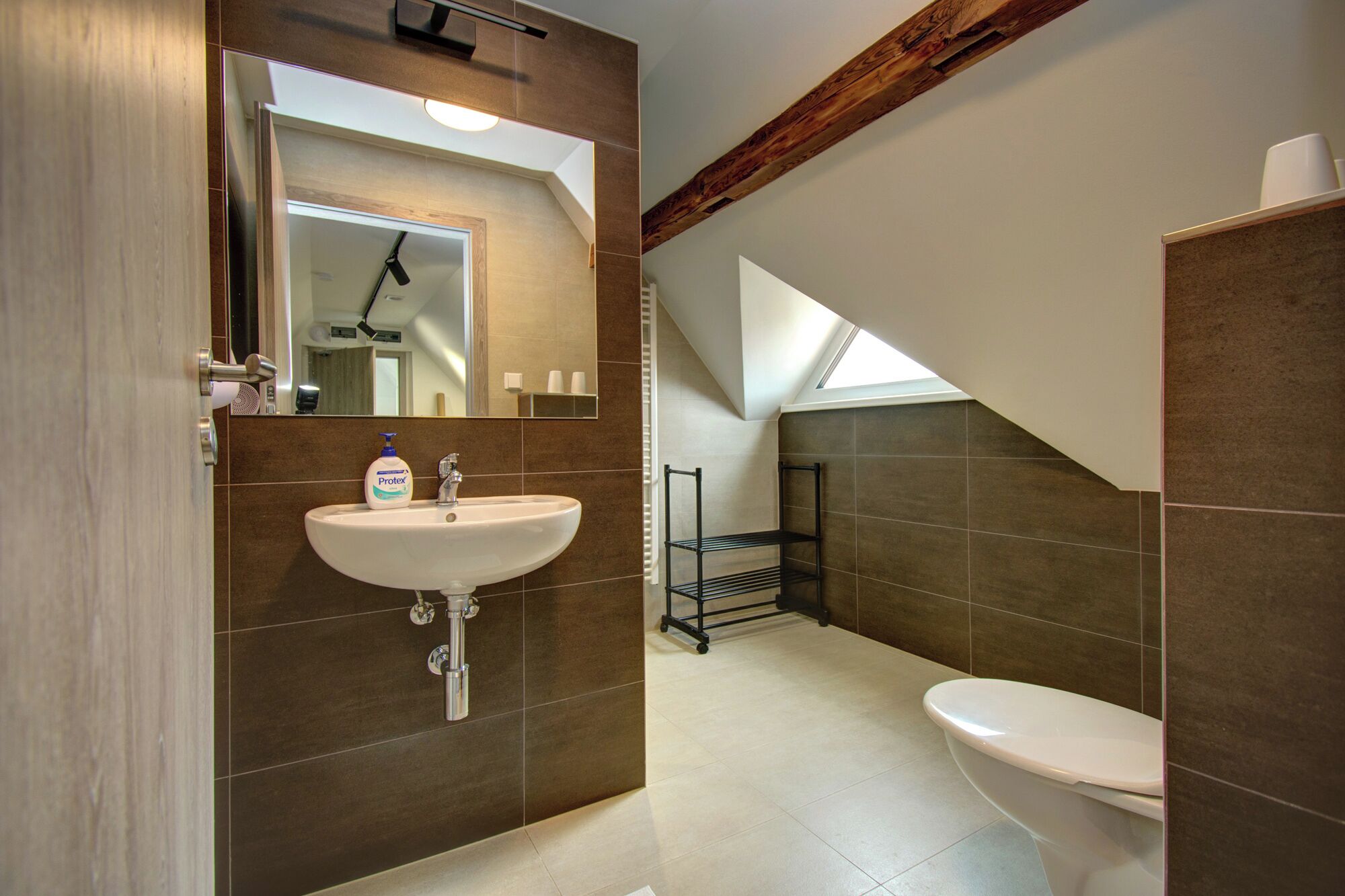 Une salle de bain moderne avec un lavabo blanc, un miroir et des toilettes. Une lucarne se trouve au-dessus des toilettes, et les murs sont recouverts de carreaux.