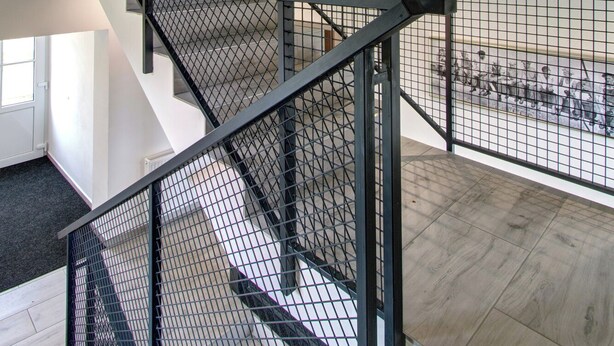 Un intérieur à plusieurs niveaux, doté d’un escalier moderne avec des rampes en métal, et offrant une vue sur une pièce où se trouve un grand fauteuil de couleur sombre et un tableau accroché au mur.