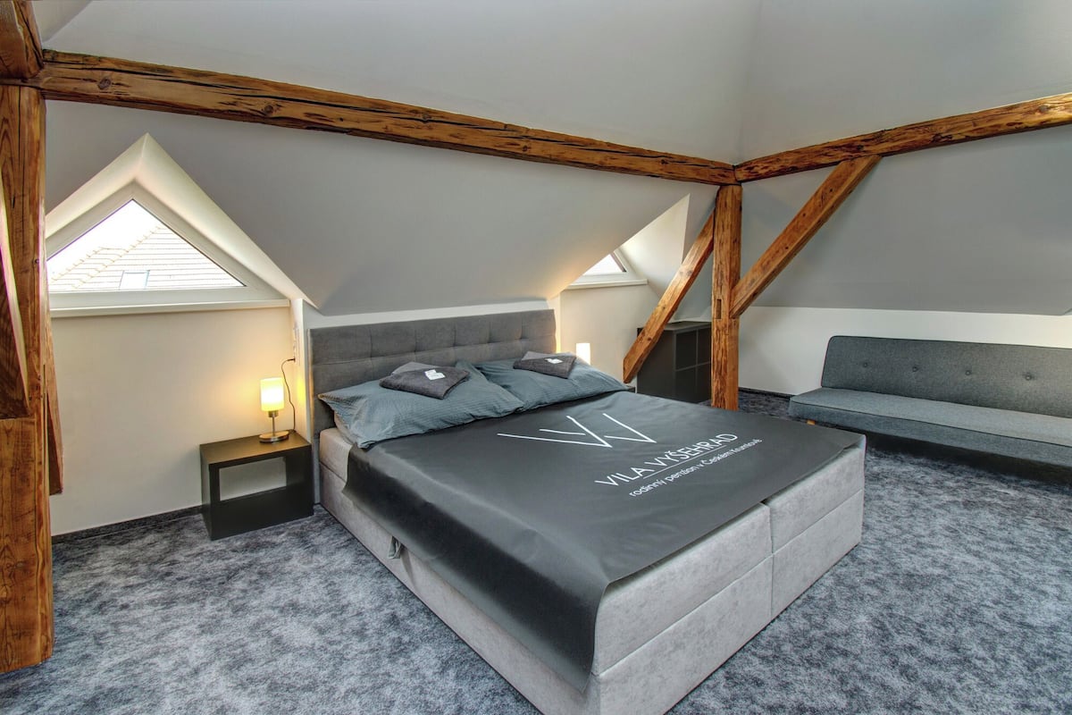 Een slaapkamer met een loft-plafond, een bed met een donkere matras en kussens, een nachtkastje met een lamp en een grijze bank.