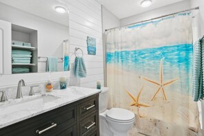 A bathroom with a shower curtain depicting a beach scene, a white toilet, a sink with a marble countertop, and a mirror above the sink.