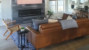 A living room with a fireplace, a brown leather sofa, a TV mounted on the wall, and a window with blinds. - Family Beach House - Pet-friendly - private heatable pool, close to the beach (Isle of Palms)