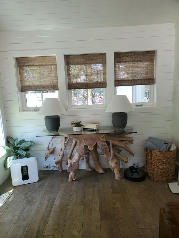 A room with a wooden floor, a large wooden table, two windows with woven blinds, a potted plant, and a small heater.