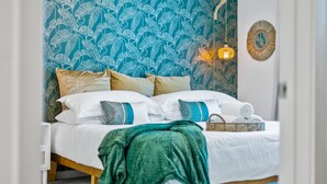 A neatly made bed with a green throw blanket, white bedding, and multiple pillows. A wicker basket with rolled towels is on the bed. The room features a blue floral wallpaper and a round mirror on the wall. - Jungle on the Square – Luxury 1BR w/ Rooftop Lounge, Pool & Gym Access (Decatur)