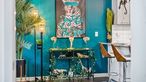 A teal-colored wall with a framed artwork of a leopard, a black console table with decorative items, and a bar area with stools. - Jungle on the Square – Luxury 1BR w/ Rooftop Lounge, Pool & Gym Access (Decatur)