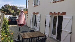 Eine Terrasse mit Tisch und Stühlen, ein geparktes Auto im Hintergrund und ein Gebäude mit weißen Fensterläden und einem Ziegeldach.