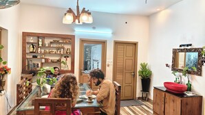 Zona pranzo con tavolo in vetro, sedie in legno e lampadario. Un mobiletto in legno con una ciotola rossa e uno specchio appeso al muro.
