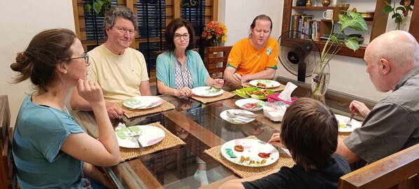 Um grupo de pessoas sentado em volta de uma mesa de vidro para refeições, com jogos americanos e pratos de comida.