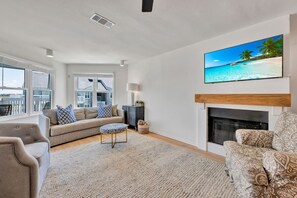 A living room with a fireplace, a large flat-screen TV, and a view of the ocean through the windows.