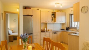 Een functionele keuken met houten kastjes, een inbouwkoelkast en een eethoek met een tafel voor twee personen.