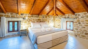 Une chambre avec un mur en pierre, des poutres en bois au plafond, un lit et deux fenêtres avec des rideaux.