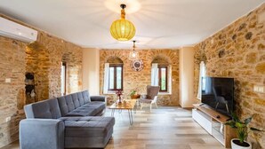 Un salon aux murs en pierre, avec un canapé gris, une table basse en bois, un téléviseur et un luminaire suspendu.