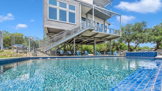 Piscina com superfície de água azul cristalina, área de deck coberta com cadeiras e um edifício de dois andares com janelas grandes e varanda.