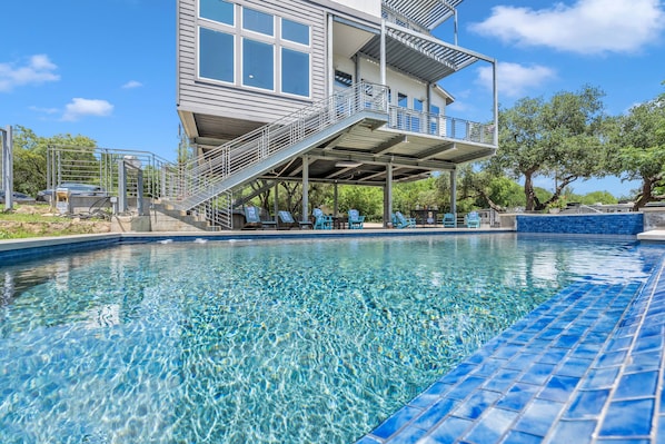 A swimming pool with a clear blue water surface, a covered deck area with chairs, and a two-story building with large windows and a balcony.