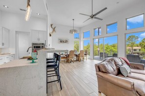 A spacious kitchen with a central island, dining area with chairs, a ceiling fan, and large windows offering a view of greenery.
