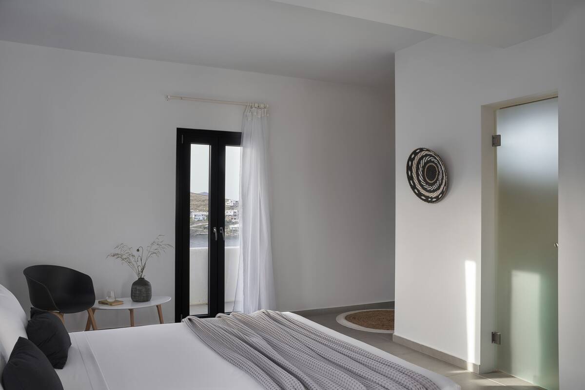 Un dormitorio moderno con cama, una silla, una mesita con un florero, una puerta y vistas al exterior.