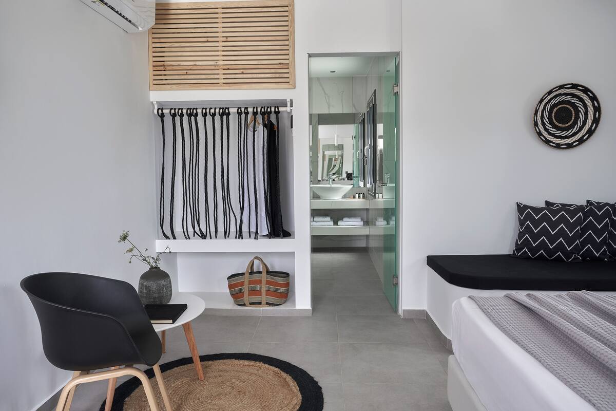 Un dormitorio moderno con una cama, una silla negra, una mesita con un florero, y un baño visible a través de una puerta de cristal.