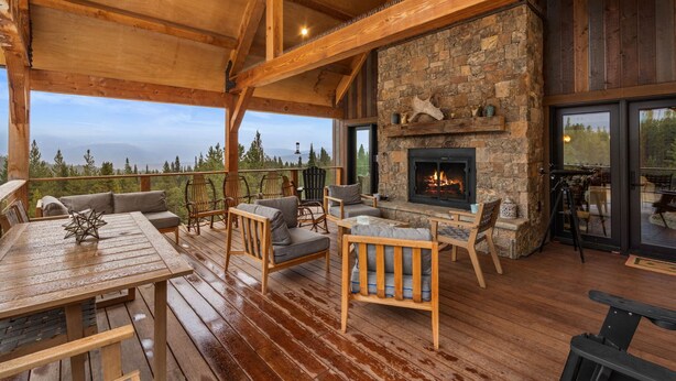 Eine Holzterrasse mit Sitzgelegenheiten im Freien, einem Kamin aus Stein und Blick auf die Berge.