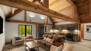 Ein geräumiges Wohnzimmer mit hoher Decke und freiliegenden Holzbalken, eine gemütliche Sitzecke mit einer Couch und Sesseln, eine Küche mit modernen Geräten und große Fenster mit Blick auf Grünflächen.
