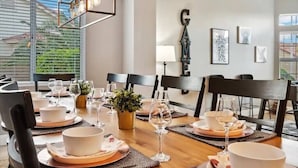 A dining area with a table set for six, including plates, bowls, and wine glasses. There are chairs around the table, a potted plant, and wall art. - Modern Majestic Home with Pool, Spa & Basketball (Mesa)
