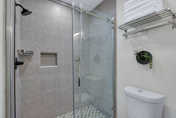 A glass-enclosed shower with a handheld showerhead, a white toilet, and a towel rack with folded towels.