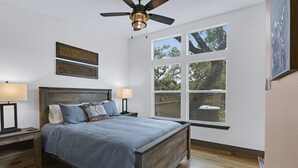 Une chambre à coucher avec un lit en bois, deux lampes de chevet, un ventilateur de plafond et de grandes fenêtres donnant sur l’extérieur.